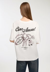 Biała, oversize'owa koszulka z czarnym nadrukiem tekstu "Ciao Amore!" oraz ilustrowanymi zawieszkami, w tym kluczami i misiem, z tyłu.