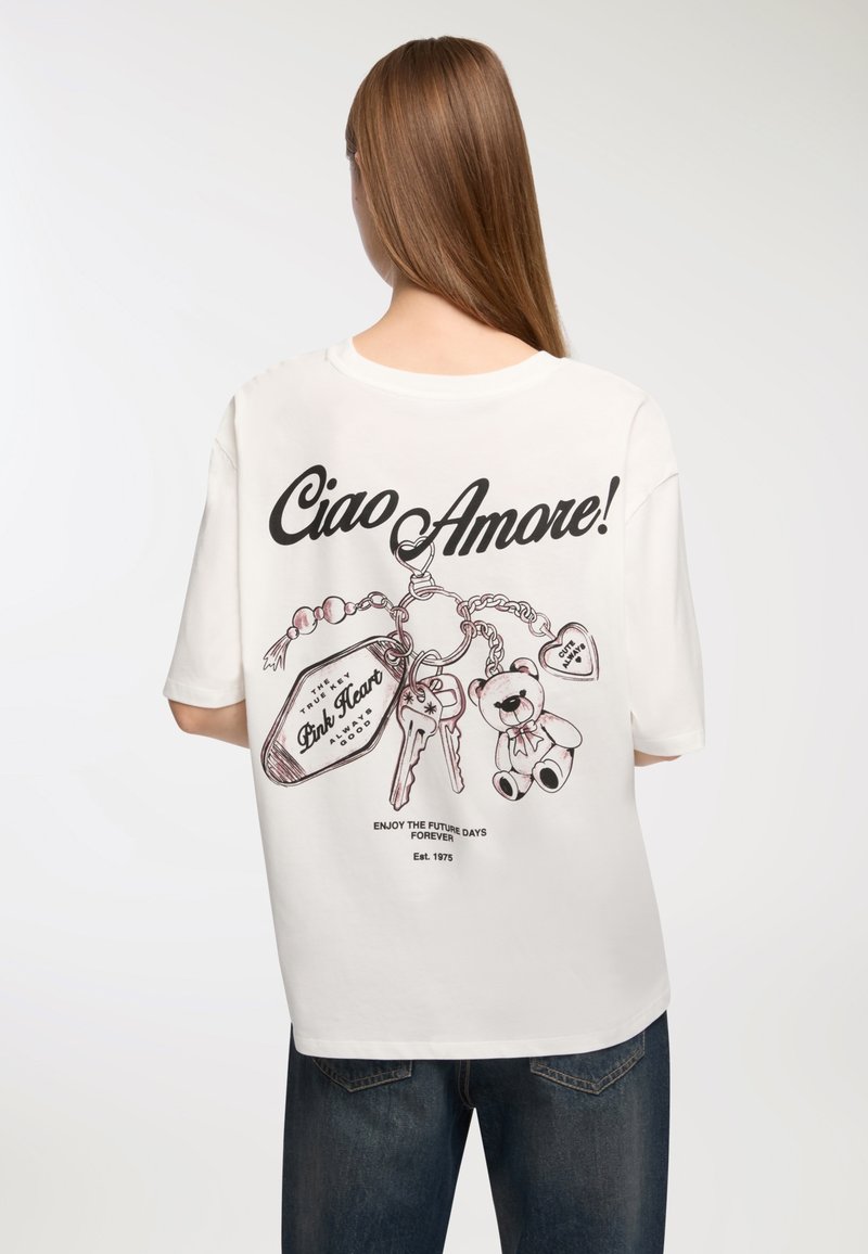 Biała, oversize'owa koszulka z czarnym nadrukiem tekstu "Ciao Amore!" oraz ilustrowanymi zawieszkami, w tym kluczami i misiem, z tyłu.