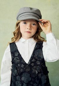 Das Kind trägt ein weißes Button-Up-Hemd unter einem schwarzen, floral bestickten Kleid, kombiniert mit einer grauen Mütze. Der Hintergrund ist sanft grün.