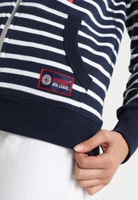 Sudadera con cremallera a rayas en azul marino y blanco con un bolsillo frontal, puños acanalados y una etiqueta roja de Superdry en la parte inferior izquierda. Textura de tela suave.