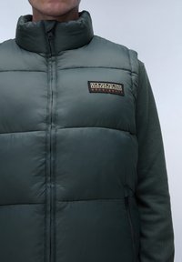 Grön vadderad väst med hög krage, prydd med en svart och orange logotypficka, dragkedjeöppning och texturerat tygdetaljer.