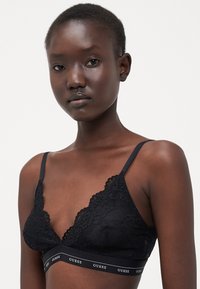 Bralette in pizzo nero con design a scallop, spalline regolabili e una fascia elastica con il logo "GUESS". Tessuto morbido.