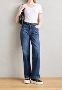 T-shirt blanc à manches courtes, jeans droits bleus, ceinture noire, sac noir et baskets noires avec semelles blanches, sur fond neutre.