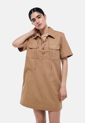 Femme aux cheveux foncés portant une robe beige à manches courtes avec un laçage sur le devant et deux poches poitrine, debout devant un fond uni.
