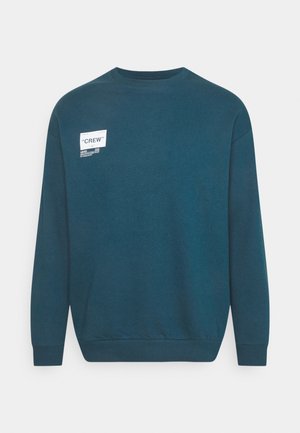 Sweatshirt en col rond teal en mélange de coton, avec une étiquette blanche sur le devant portant le texte « CREW » et des détails de marque supplémentaires.