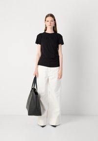 T-shirt in cotone nero con logo frontale, abbinato a pantaloni bianchi a gamba larga. Il modello tiene in mano una borsa tote in pelle nera, in piedi contro uno sfondo neutro.