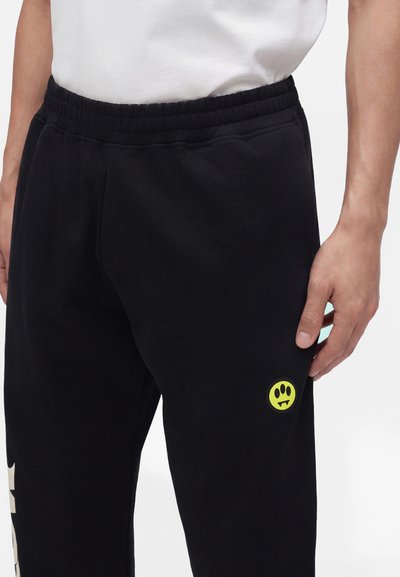 Homme portant un pantalon de survêtement noir avec une taille élastique et un petit logo jaune représentant un smiley avec des crocs sur la cuisse gauche.