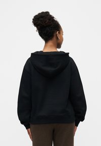 Femme aux cheveux bouclés attachés en chignon, de dos, portant un sweat à capuche noir et un pantalon marron, sur un fond blanc uni.