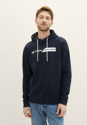 Granatowy hoodie wykonany z miękkiego materiału, z białym logo, sznurkami i wzorzystą wewnętrzną częścią kaptura. Dopasowany krój i długie rękawy.