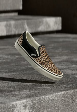 Αθλητικό παπούτσι slip-on με λεοπάρ σχέδιο και μαύρες πλευρές, αιωρούμενο πάνω από υφασμένα γκρι τσιμεντένια σκαλοπάτια.