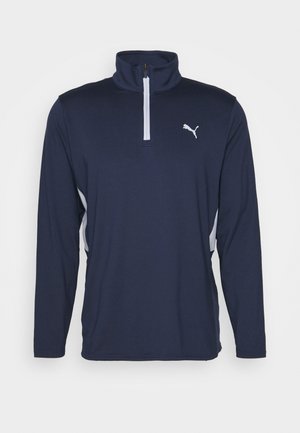 Haut de sport Puma bleu marine à manches longues, avec un col montant, une courte fermeture éclair blanche et des empiècements latéraux blancs.