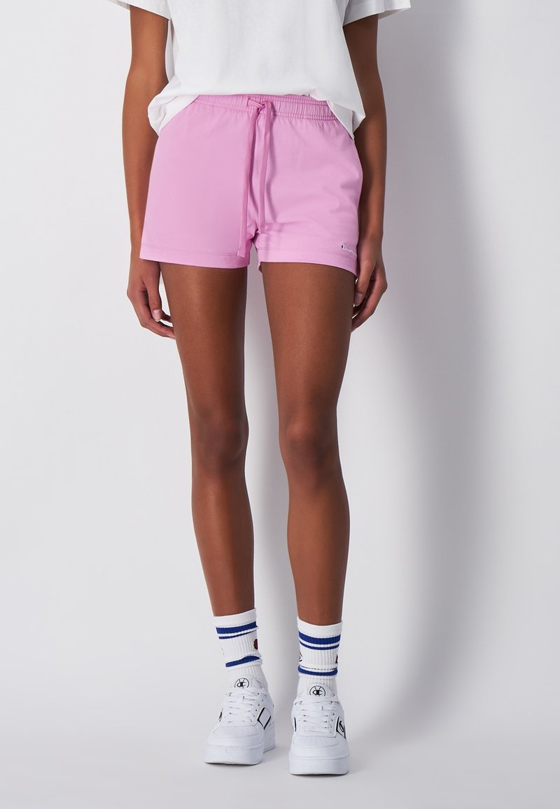 Růžové sportovní šortky s elastickým pasem a šňůrkou, s hladkým povrchem. V kombinaci s bílými teniskami a pruhovanými ponožkami.