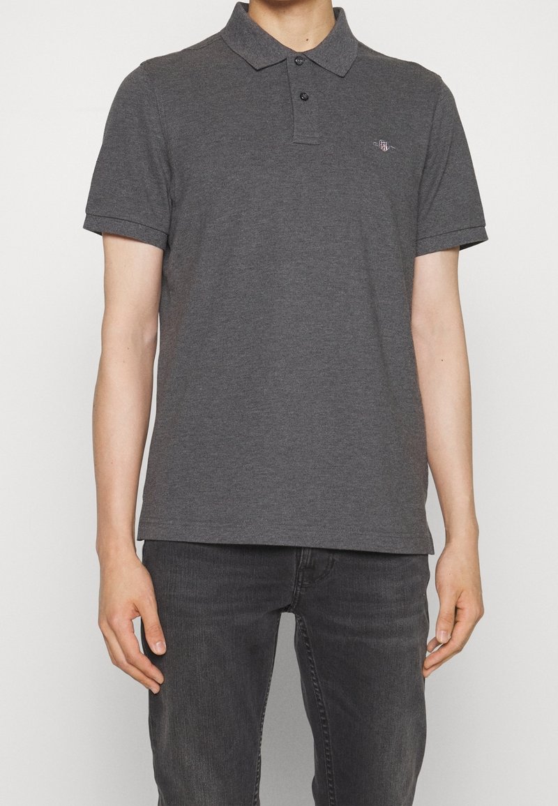 Homme portant un polo manches courtes gris foncé avec deux boutons et un petit logo brodé sur le côté gauche de la poitrine, associé à un jean gris foncé.