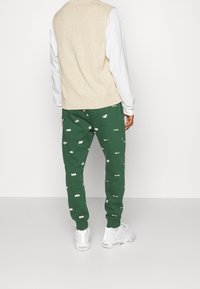 Beige s sleeveless tröja över en vit långärmad skjorta, matchad med gröna joggarbyxor med vita logotyper, och vita träningsskor.