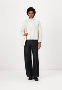 Cropped light beige jacket with blue branding, paired with dark wide-leg jeans and black shoes, accessorized with a beige handbag.