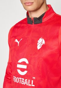 Rød sportsjakke med glidelås krage, med svarte detaljer, et teksturert mønster og hvite logoer av Puma og Fotball, plassert på forsiden.