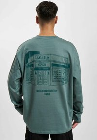 Long-sleeve green cotton sweatshirt featuring a graphic of a shop with "OPEN" text. Back displays "WORKATION COLLECTION F/W23" detail.