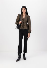 Blouse vert olive avec des manches bouffantes et des couches froncées, associée à des flares ajustées noires et des bottes noires à bout pointu, tenant un sac noir.