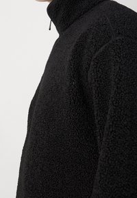 Giacca nera in pile con cerniera, caratterizzata da un colletto alto, superficie strutturata e dettagli di cuciture minime lungo le spalle e le cuciture.