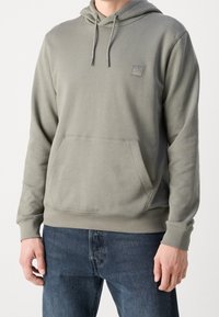 Homme portant un sweat à capuche vert olive clair avec une poche avant et des cordons, associé à un jean bleu foncé.