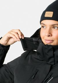Black outdoor jacket with high collar, featuring Velcro fastener, zippered pockets, and a soft texture. Accented with a leather tag on the beanie.