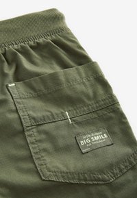 Shorts en tissu vert olive avec poche, taille élastique et une étiquette cousue sur la poche portant l'inscription "BIG SMILE Fun Times Await".