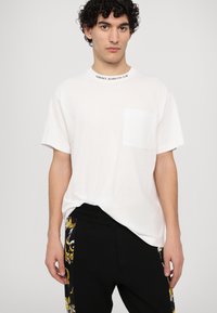 Balts kokvilnas T-krekls ar apaļu izgriezumu, ar priekšējo kabatu un izšūtu "Versace Jeans Couture" uz apkakles.