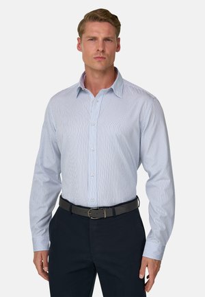 Homme portant une chemise habillée rayée verticalement bleu clair et blanche, rentrée dans un pantalon foncé avec une ceinture noire, debout devant un fond uni.
