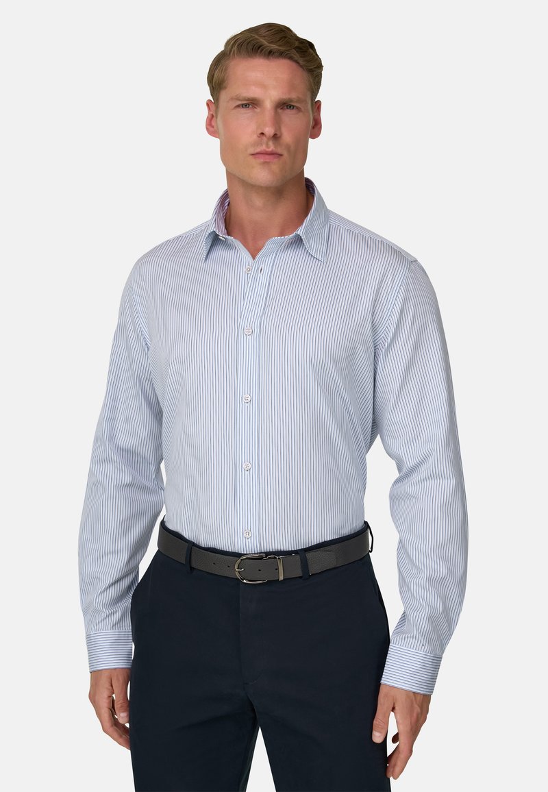 Homme portant une chemise habillée rayée verticalement bleu clair et blanche, rentrée dans un pantalon foncé avec une ceinture noire, debout devant un fond uni.