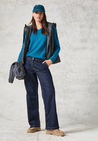 Dunkelblaue weite Jeans, türkisfarbener Langarm-Top, graue Weste und beige Schuhe. Die Mütze ist türkis; hält eine graue Jacke. Einfaches, lässiges Design.