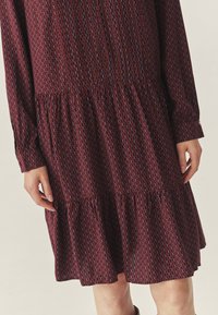 Tumepruun mustriga kleit pika varrukaga, millel on kogutud vöökoht ja laienev seelik. Disain sisaldab geomeetrilisi kujundeid sinises ja punases.