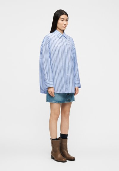Γυναίκα που φοράει υπερμεγέθη μπλε ριγωτό πουκάμισο με κουμπιά, μίνι φούστα από τζιν, μαύρες κάλτσες και καφέ μπότες μέχρι τη μέση της γάμπας, στέκεται μπροστά σε λευκό φόντο.