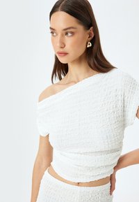 Biały, teksturowany crop top o fasonie bez ramiączek. Posiada zmarszczoną tkaninę i dopasowany krój, połączony z gładkim dekoltem.