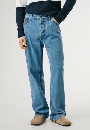 Helles blaues Denim-Jeans mit geradem Bein, ausgestattet mit einem Knopfverschluss, einer aufgesetzten Tasche und subtiler, ausgebleichter Textur. Getragen mit beigen Schuhen.