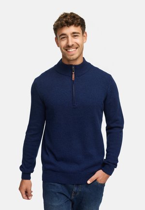 Marineblauer gestrickter Pullover mit hohem Kragen, halbem Reißverschluss und gerippten Bündchen und Saum, getragen über einer blauen Jeans.