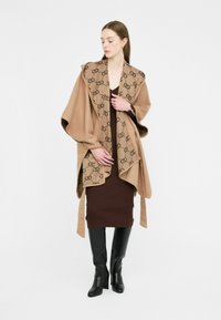 Poncho beige con un interno marrone a fantasia, caratterizzato da un design asimmetrico. Il modello indossa un vestito marrone attillato e stivali neri al ginocchio.