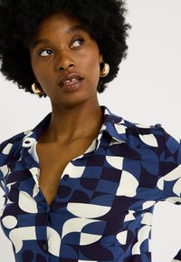 Chemise à boutons avec un motif géométrique bleu marine et blanc. Le matériau est doux avec un léger brillant, dotée d'un col et d'une coupe ajustée.