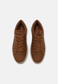 Sneakers din piele maro cu vârfuri rotunjite, șase găuri pentru șireturi și o suprafață texturată. Au o talpă din cauciuc alb contrastant și detalii perforate.