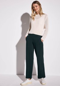Maglione lavorato a maglia di un beige chiaro con maniche lunghe, abbinato a pantaloni larghi verde scuro con tasche laterali. Indossato con sneakers bianche.