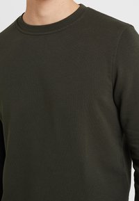 Maglietta a maniche lunghe verde scuro realizzata in morbido cotone. Presenta un collo rotondo e dettagli di cucitura minimalisti sul colletto e sulle maniche.