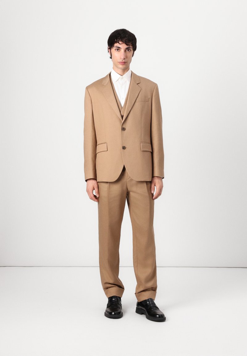 Beige tailored suit with a single-breasted jacket, notch lapels, two front pockets, and matching trousers. Worn with a white shirt and black shoes.
