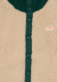 Veste en polaire beige avec des boutons-pression et un col vert foncé, comportant une petite étiquette "TINY" en rouge sur la poitrine.
