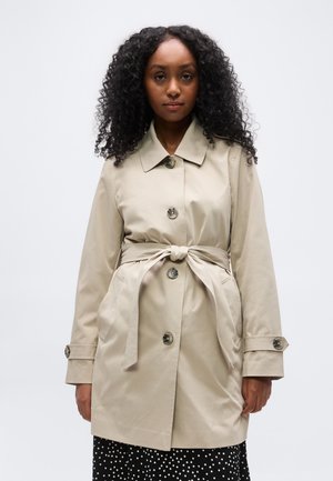 Γυναίκα με σγουρά μαλλιά, ντυμένη με μπεζ καμπαρντίνα με ζώνη πάνω από μια μαύρη και λευκή φούστα με πουά, στέκεται μπροστά σε ένα απλό φωτεινό φόντο.