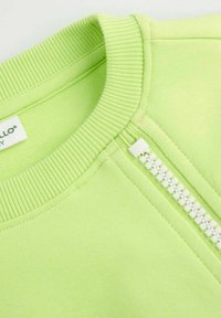 Jasnozielony sweter z ribbowanym kołnierzem, z białym zamkiem po jednej stronie i gładką fakturą materiału. Widoczna metka wewnątrz.