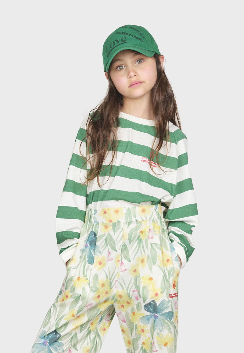 Camicia a maniche lunghe a righe verdi e bianche abbinata a pantaloni con stampa floreale, caratterizzati da una combinazione di colori pastello. Cappello verde con dettagli testuali.