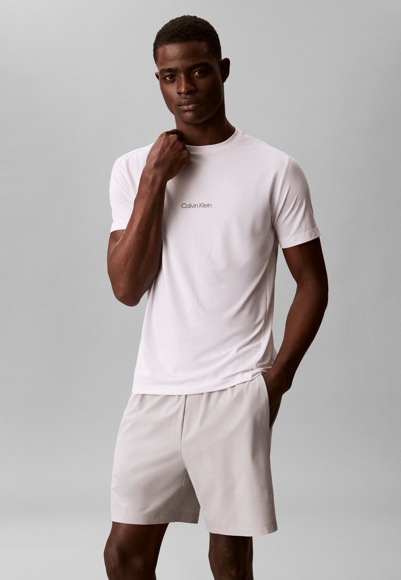 Άνδρας που φοράει λευκό t-shirt Calvin Klein και ανοιχτόχρωμα σορτς στέκεται μπροστά σε απλό γκρι φόντο με το ένα χέρι στην τσέπη.