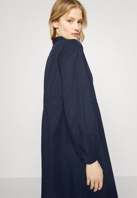 Vestido azul-marinho com colarinho alto, mangas bufantes longas e cintura franzida. Apresenta um design descontraído e um tecido leve.