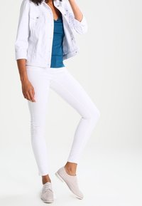 Vit denimjacka, turkos linne, vita skinny jeans och ljusgrå snörskor. Släta texturer och figursydd design med subtila detaljer.