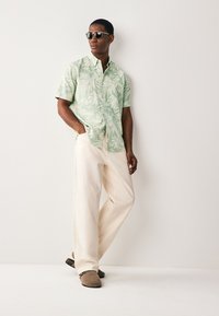 Camicia verde a maniche corte con stampa di foglie di palma, pantaloni beige a gamba larga, scarpe slip-on grigie e occhiali da sole. Outfit semplice e casual.