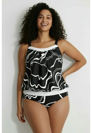 Uśmiechnięta kobieta z kręconymi włosami w czarno-białym tankini z abstrakcyjnym wzorem, stojąca na białym tle.